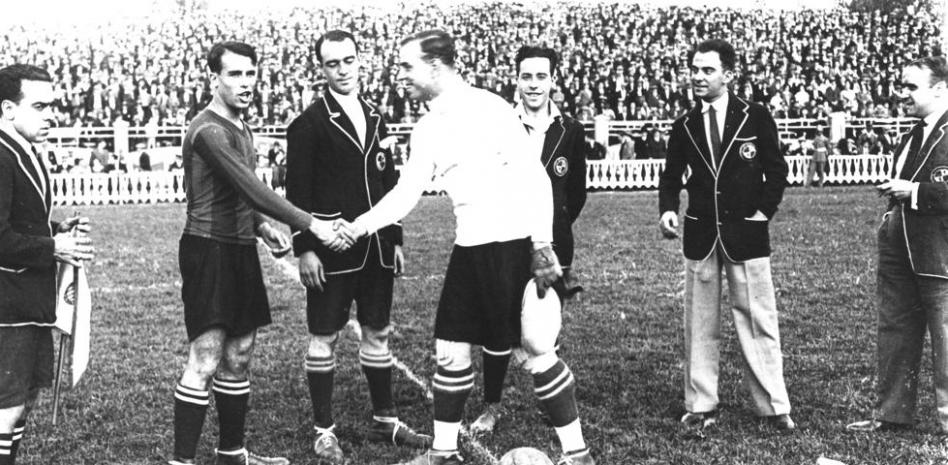 Dos capitanes de equipos de fútbol, uno del FC Barcelona y otro del RCD Espanyol, se saludan en el centro del campo antes de un partido. A su alrededor, están rodeados de otros jugadores y oficiales del club, con una multitud de espectadores llenando las gradas al fondo.